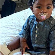 Salish joined the competition — help win amazing prizes! baby, child, pacifier, headband, bow, curly_hair, denim_dress, white_shoes, bed, bedspread, indoors, sitting, cute, infant, person, portrait, clothing, footwear, young_child, closeup