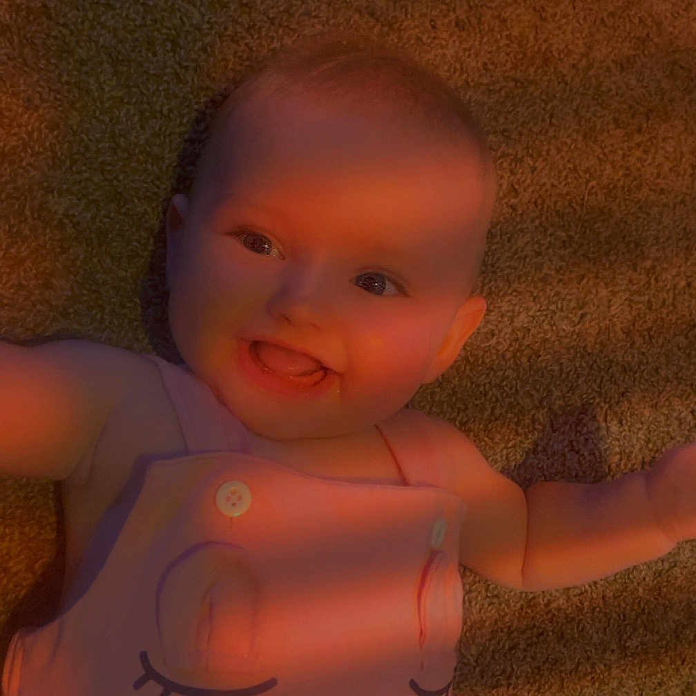 Kora is registered to the contest to win money with this photo: apparel, baby, carpet, cheerful, child, cute, eyes, face, happy, indoor, infancy, infant, laying, person, pink_dress, portrait, shadow, smiling, sunlight, texture