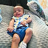 baby, smiling, indoor, pillow, blue_shorts, colorful_shirt, happy, couch, child, person, footwear, white_shoes, plush, relaxing, tongue_out, seated, texture, cozy, furniture, home