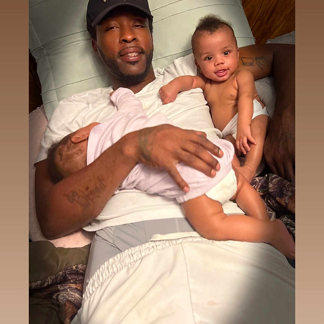 My'Drevious Bell is registered to the contest to win money with this photo: man, baby, infant, child, sleeping, smiling, bed, pillow, cap, tattoo, white_shirt, white_pants, family, indoor, relaxing, skin, hand, lying_down, cozy, portrait