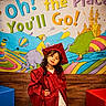 child, graduation, gown, cap, pose, colorful, mural, illustration, hot_air_balloon, animal, celebration, smile, curly_hair, confident, indoor, wooden_wall, red, blue, yellow, green