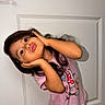 Leiza is registered to the contest to win money with this photo: child, girl, pose, pink_shirt, long_hair, door, indoor, cute, expression, playful, hands_on_face, standing, portrait, young_child, casual_clothing, fun, smile, person, human, wall