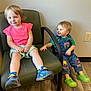 Morgan is registered to the contest to win money with this photo: toddler, child, smiling, indoor, chair, green_chair, denim_overalls, pink_shirt, blue_shoes, green_shoes, wooden_floor, wall_outlet, light_wall, window, happy, cute, playful, seated, floor, person