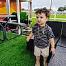 backyard, building, chair, child, curly_hair, daylight, fence, furniture, gold_chain, grass, outdoor, patio, playful, portrait, shoes, shorts, standing, striped_shirt, table, toddler
