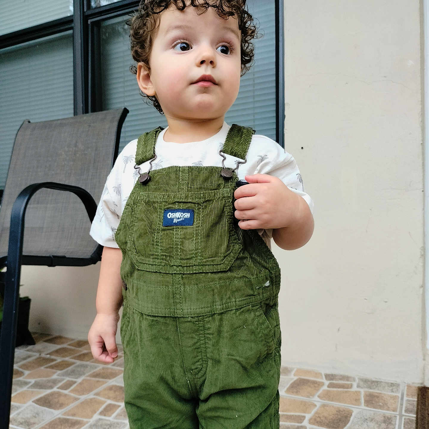 Enzo Calero Cepeda is registered to the contest to win money with this photo: child, toddler, curly_hair, portrait, standing, outdoor, patio, overalls, green_clothing, corduroy, white_shirt, looking_away, neutral_expression, face, eyes, window, chair, tiled_floor, daylight, casual_photo