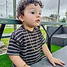 Enzo Calero Cepeda joined the competition — help win amazing prizes! child, toddler, curly_hair, portrait, outdoor, table, picnic_table, striped_shirt, gold_chain, bracelet, sitting, standing, face, eyes, hands, grass, patio, chairs, utility_poles, overcast_sky