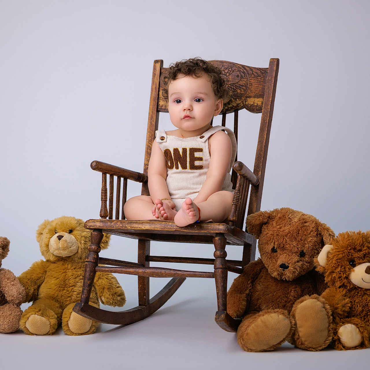 Enzo Calero Cepeda is registered to the contest to win money with this photo: baby, child, toddler, sitting, rocking_chair, wooden_chair, teddy_bears, stuffed_animals, portrait, studio, indoors, white_background, curly_hair, onesie, bare_feet, serious_expression, infant, furniture, soft_toys, centered_composition