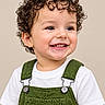 toddler, child, curly_hair, smiling, teeth, portrait, studio, close_up, overalls, green_clothing, white_shirt, happy, face, eyes, hair, baby, person, indoor, beige_background, fashion