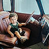 toddler, child, baby, vintage_car, car_interior, driver_seat, steering_wheel, dashboard, leather_seat, chrome, curly_hair, portrait, sitting, looking_at_camera, green_outfit, shoes, automobile, indoor, photography, cute