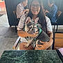 woman, dog, french_bulldog, puppy, glasses, table, green_marble, chair, outdoor, people, elderly, eating, casual_clothing, smiling, happy, seated, restaurant, daylight, cement_floor, fence