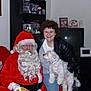 person, santa_claus, woman, dog, indoor, glasses, leather_jacket, white_dog, television, bookshelf, holiday_decorations, snowman_figurine, smile, curly_hair, christmas, seated, red_suit, white_beard, festive