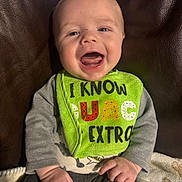 Dean is registered to the contest to win money with this photo: baby, smiling, bib, green_bib, clothing, sitting, infant, happy, mouth_open, hand, fingers, gray_sleeves, dark_pants, blanket, couch, leather, indoor, face, person, cute
