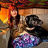 person, dog, black_dog, couch, pajamas, indoor, wood_paneling, tapestry, colorful, red_hair, relaxed, seated, pet, companion, casual_clothing, animal, living_room, fabric, artistic_background, cozy