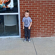Benny is registered to the contest to win money with this photo: child, boy, brick_wall, sidewalk, window, glass, poster, smiling, casual_clothing, tshirt, sneakers, outdoor, daylight, concrete, person, young, happy, portrait, urban, cute