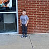 child, boy, brick_wall, sidewalk, window, glass, poster, smiling, casual_clothing, tshirt, sneakers, outdoor, daylight, concrete, person, young, happy, portrait, urban, cute