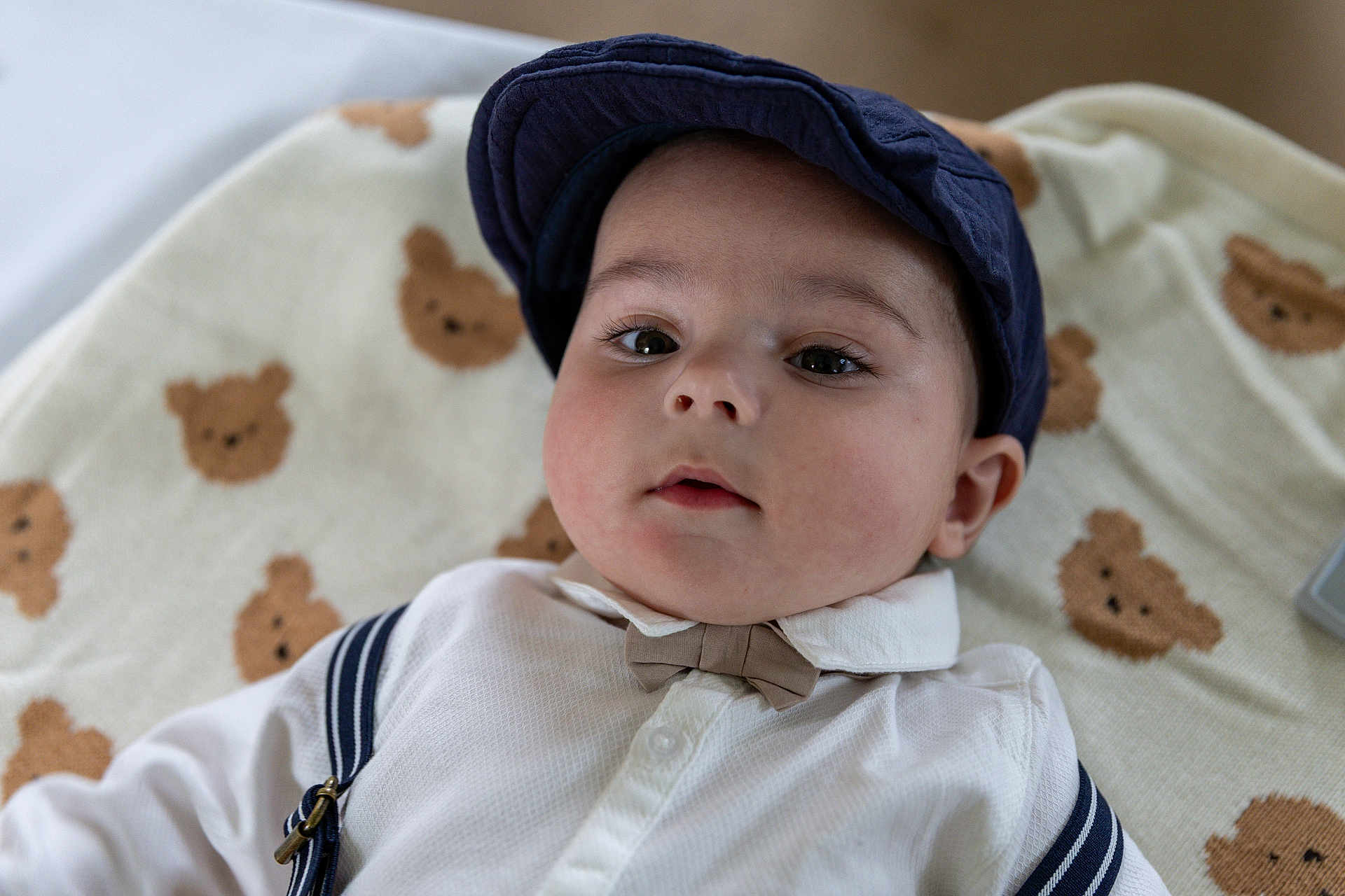 Nicolas joined the competition — help win amazing prizes! baby, child, face, cap, hat, bow_tie, shirt, suspenders, blanket, teddy_bear_pattern, infant, cute, portrait, lying_down, indoor, clothing, young_child, expression, closeup, headwear