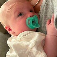 Jackson is registered to the contest to win money with this photo: baby, pacifier, infant, close_up, person, adult, hand, clothing, necklace, skin, face, ear, white_shirt, gray_shirt, cuddle, comfort, indoor, portrait, young_child, holding