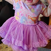 Kiara is registered to the contest to win money with this photo: baby, child, toddler, standing, smiling, bow, headband, tutu, dress, purple, colorful, patterned_clothing, barefoot, portrait, indoors, adult, tattoo, carpet, furniture, happy