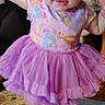 baby, child, toddler, standing, smiling, bow, headband, tutu, dress, purple, colorful, patterned_clothing, barefoot, portrait, indoors, adult, tattoo, carpet, furniture, happy