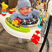 Nathaniel is registered to the contest to win money with this photo: baby, child, walker, toy, smiling, blue_hat, indoor, floor, furniture, baby_clothing, colorful_toys, happy, person, seat, infant, play, wood_floor, couch, baby_walker, cute