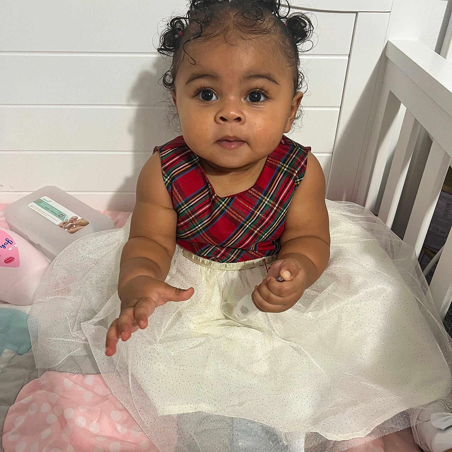 Kaylani is registered to the contest to win money with this photo: baby, child, infant, girl, curly_hair, plaid, dress, tulle_skirt, crib, blanket, wipes, soft_toys, indoor, cute, smiling, person, portrait, sitting, hand, baby_clothing