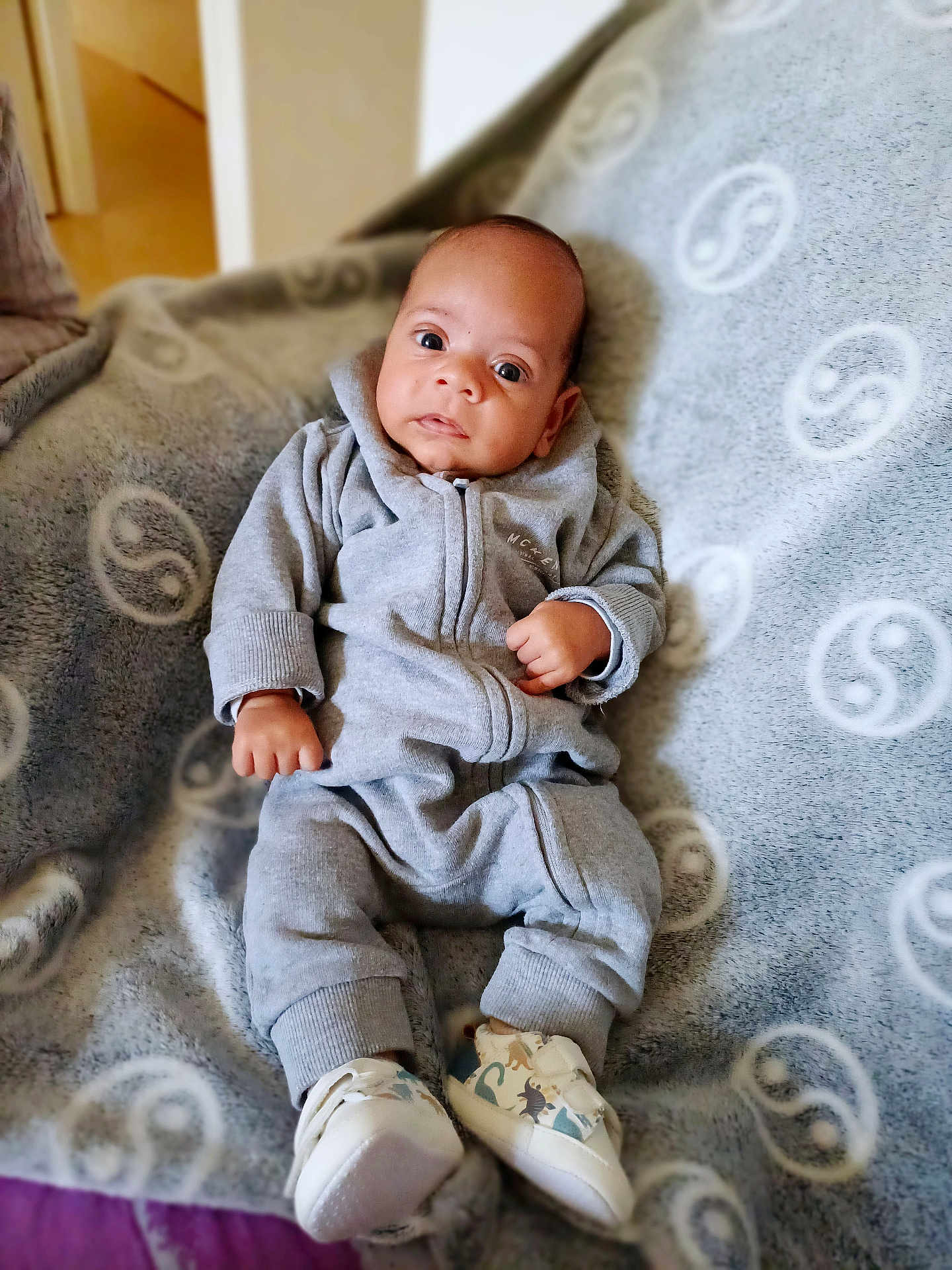 Tyano a rejoint le concours — aidez-le/la à gagner de superbes lots ! baby, infant, onesie, grey_clothing, blanket, soft_texture, dinosaur_shoes, footwear, indoor, curious_expression, lying_down, cozy, child, cute, small_hands, patterned_blanket, comfortable, portrait, wide_eyes, young_child