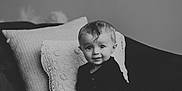 Oaklee is registered to the contest to win money with this photo: baby, child, infant, couch, pillow, black_and_white, portrait, smile, curly_hair, indoor, cozy, seated, cute, young, person, home, soft_light, looking_at_camera, clothing, footwear