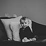 baby, child, infant, couch, pillow, black_and_white, portrait, smile, curly_hair, indoor, cozy, seated, cute, young, person, home, soft_light, looking_at_camera, clothing, footwear