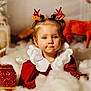 toddler, child, holiday, christmas, festive, reindeer_antlers, red_clothing, white_collar, gift_box, fluffy_decorations, pine_cone, portrait, indoor, cute, smiling, blue_eyes, holiday_decor, soft_lighting, blurry_background, festive_headband