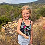 child, girl, freckles, braids, smile, insect, hand, nature, outdoor, rocks, path, grass, hill, cloudy_sky, summer_clothing, skirt, tank_top, sandals, person, happy