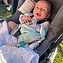 baby, stroller, blanket, smiling, outdoor, grass, person, toy, child, clothing, seatbelt, sunlight, happy, infant, face, hand, fabric, footwear, leg, casual