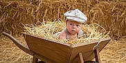Raphaël a rejoint le concours — aidez-le/la à gagner de superbes lots ! baby, wheelbarrow, straw, hay, barn, vintage_cap, child, wooden, rustic, curious, indoors, face, portrait, agriculture, farm, cute, infant, sitting, natural_light, costume