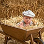 baby, wheelbarrow, straw, hay, barn, vintage_cap, child, wooden, rustic, curious, indoors, face, portrait, agriculture, farm, cute, infant, sitting, natural_light, costume