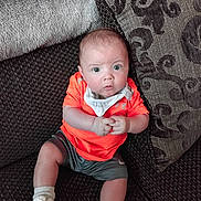 Grayson-Jo is registered to the contest to win money with this photo: baby, infant, child, couch, pillow, orange_shirt, white_shoes, socks, shorts, indoor, curious, hands, seated, looking_up, home, fabric, patterned_pillow, soft_texture, casual_clothing, portrait
