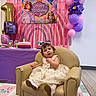 toddler, birthday_party, balloons, cake, birthday_candle, armchair, dress, child, indoor, celebration, banner, princess_theme, purple_tablecloth, floor_mat, party_decorations, smiling, young_child, festive, happy, seated