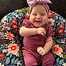 baby, infant, smiling, purple_clothing, headband, bow, floral_cushion, happy, sitting, cute, child, indoors, portrait, comfort, face, person, small_hands, cozy, pattern, colorful