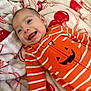 baby, infant, smiling, orange_clothing, striped_clothing, jack_o_lantern, blanket, soft_texture, red_and_white, happy, lying_down, cute, portrait, child, young, cozy, indoors, warm, face, expressive