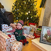 Xavier-James is registered to the contest to win money with this photo: baby, candy, chair, christmas_tree, decorations, elf_costume, festive, floor, framed_photo, hand, holiday, indoors, living_room, person, presents, socks, sofa, wallpaper, wooden_floor, wrapped_gifts