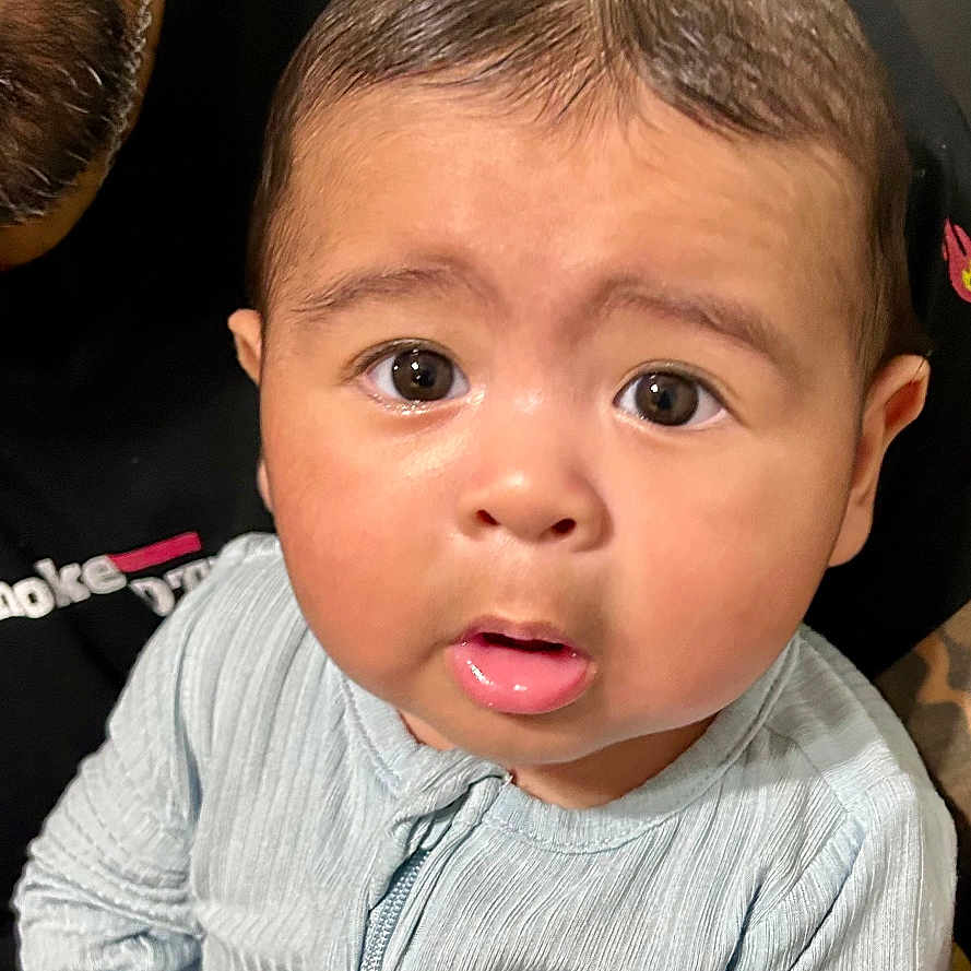 Leo Santana is registered to the contest to win money with this photo: adult, baby, blue_outfit, child, clothing, curious, cute, expression, face, hands, holding_toy, indoors, infant, lap, person, portrait, seated, skin, tattoo, wide_eyes