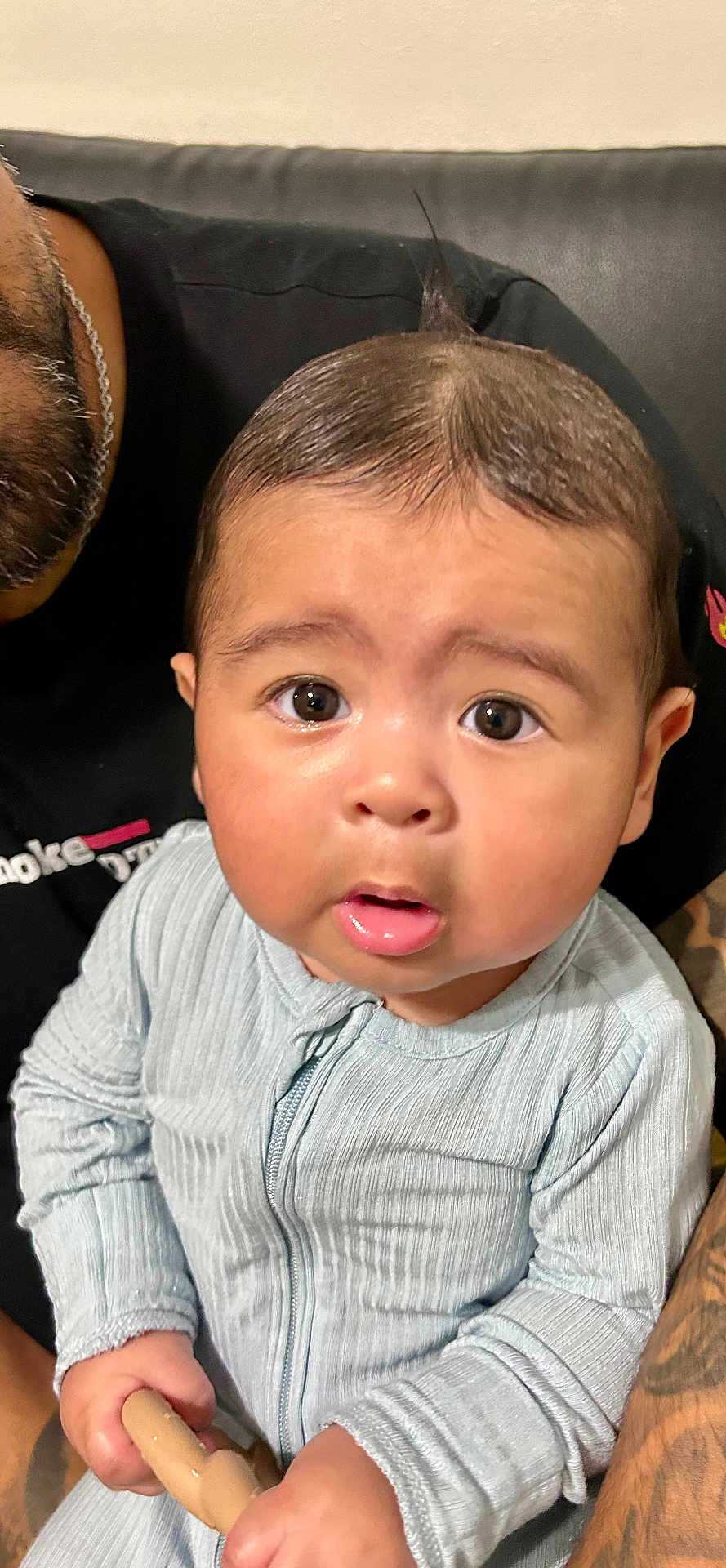 Leo Santana is registered to the contest to win money with this photo: baby, child, infant, cute, wide_eyes, curious, holding_toy, tattoo, adult, lap, clothing, blue_outfit, portrait, indoors, skin, face, hands, expression, person, seated