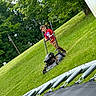 child, boy, outdoor, lawn, grass, mower, lawn_mower, trampoline, backyard, trees, greenery, smiling, standing, playful, summer, red_shirt, brown_shorts, yard_work, equipment, tilted_photo