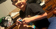 Aiden is registered to the contest to win money with this photo: child, boy, smiling, portrait, indoor, bedroom, bed, dresser, television, toy_truck, snack, wrapper, black_shirt, shorts, sitting, home, furniture, colorful, happy, casual