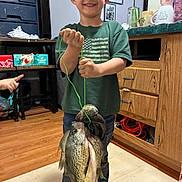 Aiden joined the competition — help win amazing prizes! child, boy, smiling, fishing, fish, catch, angler, indoor, portrait, standing, green_shirt, american_flag_shirt, jeans, fishing_line, trophy, proud, home, wood_floor, furniture, family
