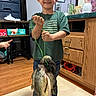 child, boy, smiling, fishing, fish, catch, angler, indoor, portrait, standing, green_shirt, american_flag_shirt, jeans, fishing_line, trophy, proud, home, wood_floor, furniture, family