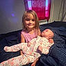 child, baby, siblings, girl, infant, portrait, smiling, sitting, bed, bedroom, floral_clothing, pink_shirt, long_hair, family, indoors, curtains, window, blanket, cozy, holding_baby