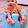 baby, infant, smiling, blue_baby_seat, orange_clothing, happy, indoors, flooring, child, cute, sitting, small_toddler, toes, face, person, home, floor, bright, expression, seat