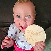 Pj is registered to the contest to win money with this photo: baby, infant, eight_months, smiling, blue_eyes, bib, shorts, hand, couch, indoor, child, person, holding, wooden_sign, happy, sitting, closeup, face, cute, portrait