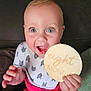 baby, infant, eight_months, smiling, blue_eyes, bib, shorts, hand, couch, indoor, child, person, holding, wooden_sign, happy, sitting, closeup, face, cute, portrait