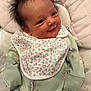 baby, smiling, face, hair, onesie, bib, floral_pattern, green_clothing, lying_down, quilted_surface, infant, cute, happy, portrait, child, newborn, soft_lighting, indoors, cozy, toddler
