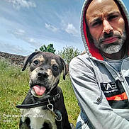 Sayan participe au concours pour gagner de l'argent avec cette photo : beard, casual, clouds, collar, daylight, dog, friendship, grass, hoodie, leisure, man, nature, outdoor, person, portrait, puppy, sky, smiling, stone_wall, tongue_out